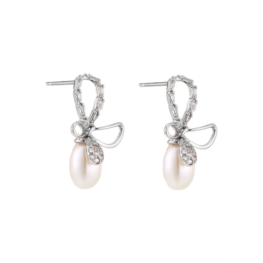 Silver bow-shaped stud earrings with baguette crystals and dangling white freshwater pearls on cream background